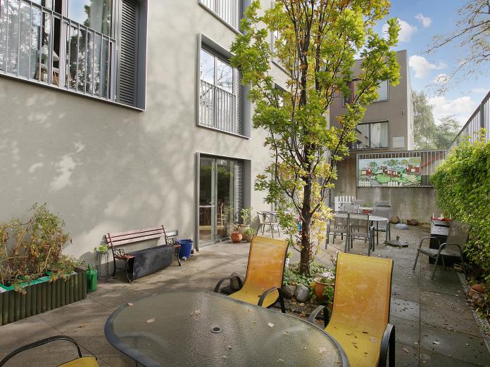 Terrasse med havemøbler, bænk, træer, højbed og buske.