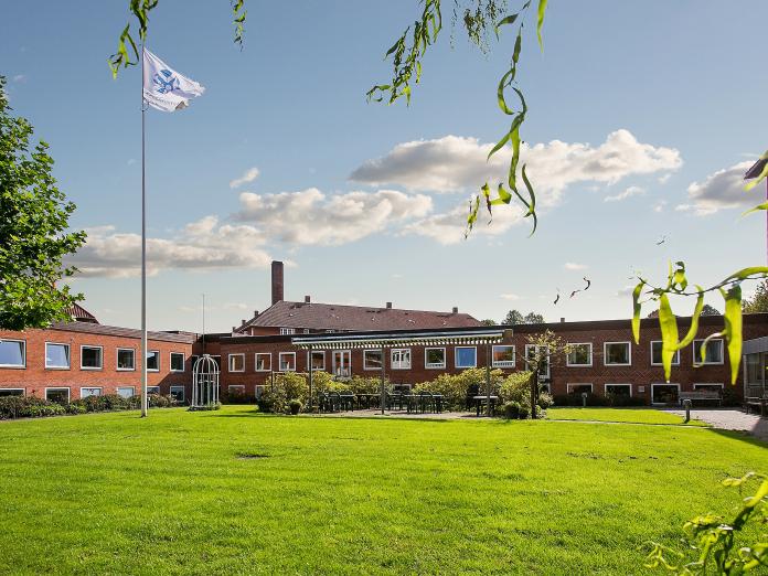 Terrasse med havemøbler og markise omkranset af en grøn græsplæne med flagstang. Bag terrassen er et lavt, rødt murstensbyggeri med hvide vinduer.
