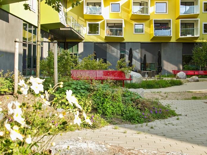 Gårdhave med grønne bede, træer flisegangstier og halvcirkelbænke. 