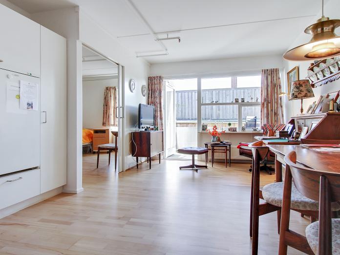 Lys stue med tekøkken og åben dør ud til altan. Stuen er indrettet med personlige møbler som spisebord med stole og lænestol, sidebord og fjernsyn.