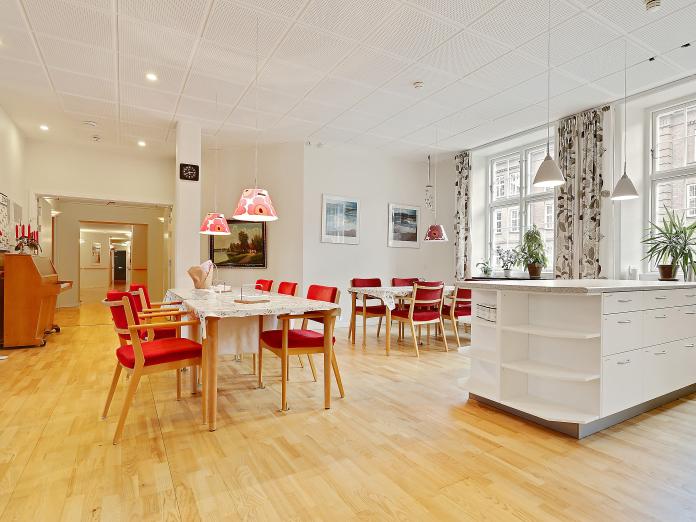 Fællesstue med klaver, bord og stole samt serveringsdisk. Stuen er lys med højt til loftet og store vinduer.