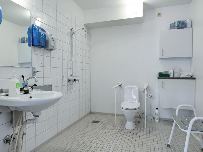 Stort, lyst flisebadeværelse med vask, spejl, toilet med armstøtter, bruser og badebænk.