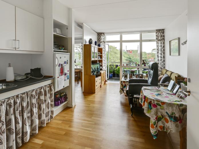 Lys, aflang stue med gulv til loft-vinduer og dør ud til altan. Stuen er indrettet med et tekøkken med vask og køleskab samt møbleret med personlige møbler.