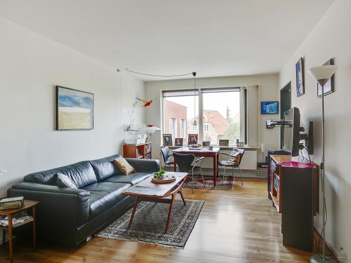 Stue med sofa, sofabord og tv-møbel. Ved vinduerne står et spisebord med tre spisebordsstole. På væggene hænger forskellige billeder. 