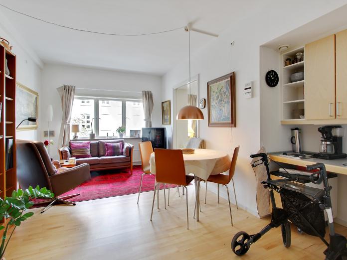 Lyst værelse med tekøkken indrettet med personlige møbler som sofa, sofabord, lænestol, fjernsyn og lille spisebord med stole omkring. 