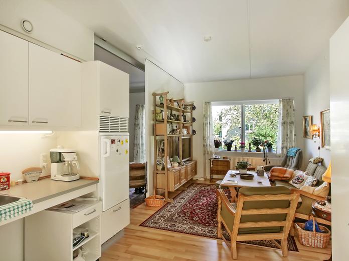 Lys stue med tekøkken og indbyggede skabe. Stuen er indrettet med personlige møbler som sofa, sofabord og lænestol samt et reolsystem og malerier på væggene.