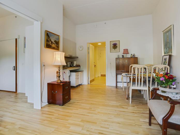 Lys stue med trægulve og tekøkken indrettet med personlige møbler som spisebord med stole samt lænestol, sideborde, lamper og malerier på væggene.