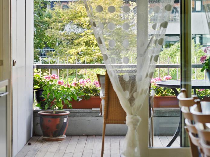 Udkig gennem terrassedør ud til en træterrasse med havemøbler, altankasser med blomster og blomster i krukker.