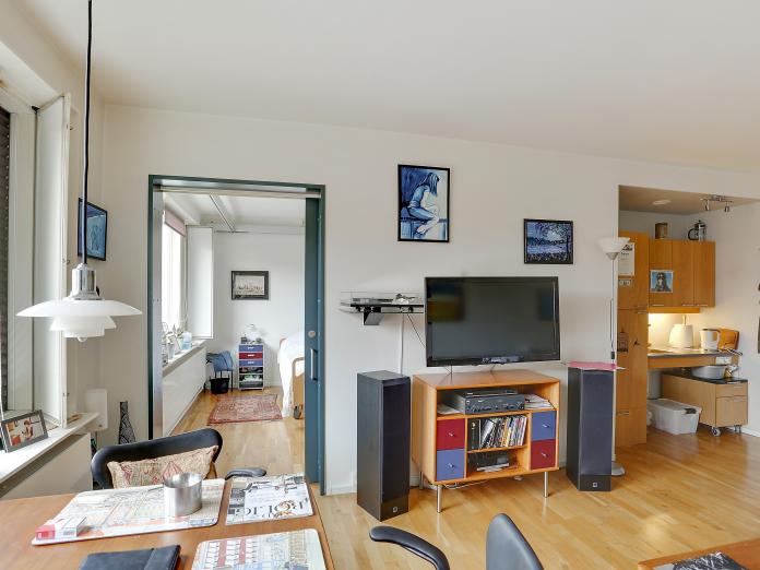 Stue med kig ind til soveværelse og skydedør imellem værelserne, et lille tekøkken med køleskab, skabslåger og vask.