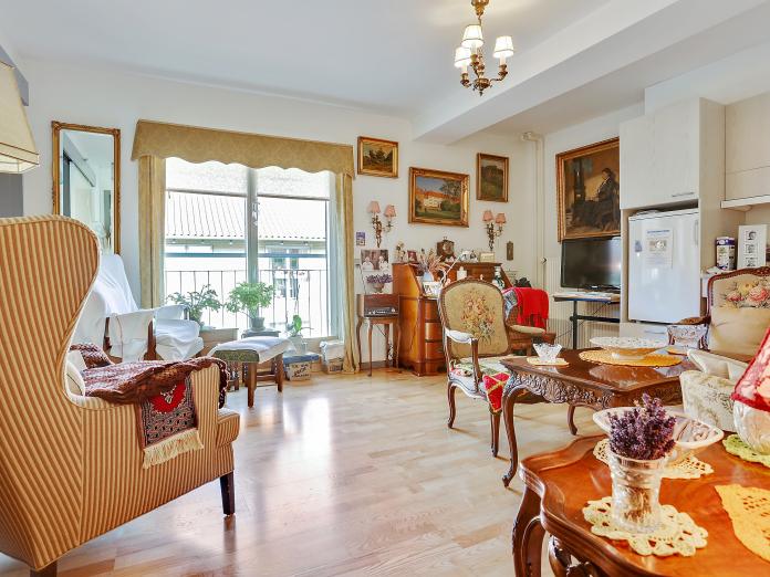 Lys stue med stort vindue og dør ud til altan. Stuen indeholder også et tekøkken i lyst træ med køleskab og vask. Derudover er stuen møbleret med personlige møbler i mørkt træ.