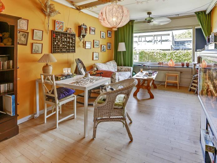 Værelse med gule vægge og stort vinduesparti. Værelset er indrettet med personlige møbler som sofa, sofabord, gyngestol, spisebord, stol og et stort akvarie.