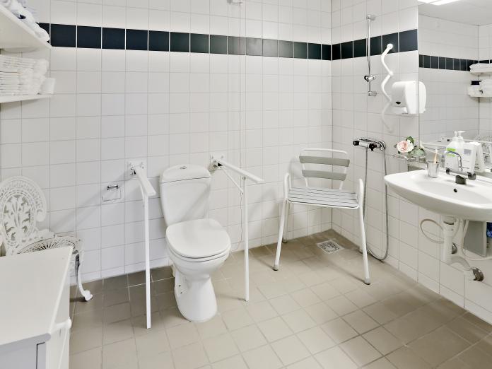 Stort, lyst flisebadeværelse med toilet, vask, bruser og hylder til personlige ting.