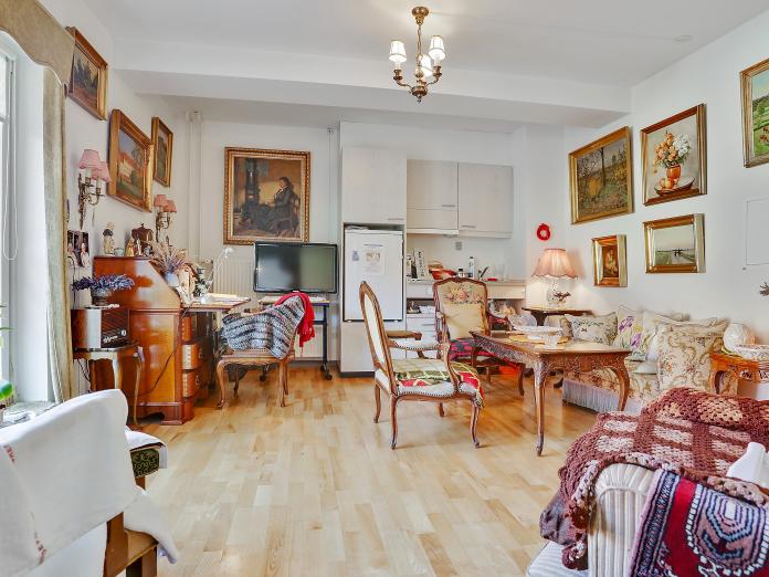 Lys stue med tekøkken i lyst træ med køleskab og vask. Stuen er indrettet med personlige møbler som sofa, lænestole, sofabord, chatol, fjernsyn og malerier på væggene.