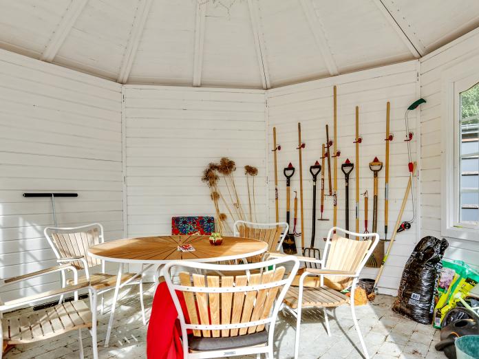 Lille tehus med et rundt havebord, fem havestole og en masse haveredskaber, der hænger på væggen. På jorden står forskelligt jord i poser, og der er også et lille vindue.