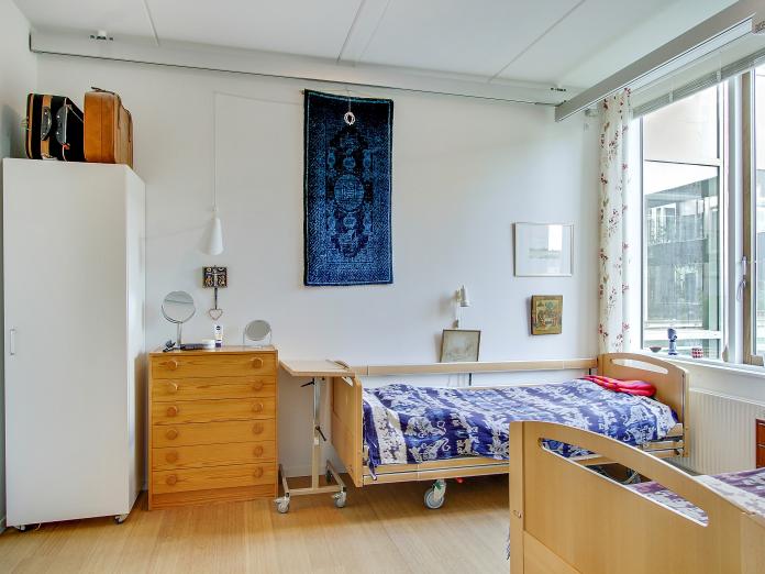 Soveværelse med to plejesenge, en lille kommode, et skab med kufferter ovenpå, billeder og vægtæppe.