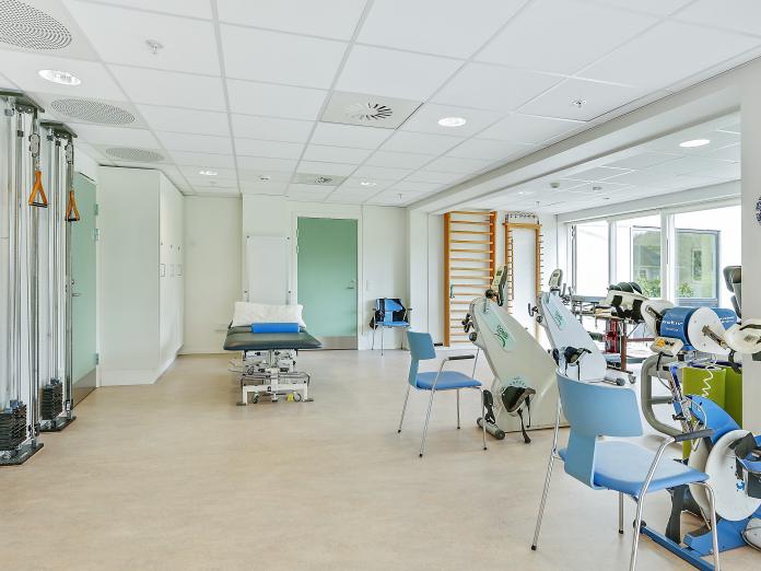 Lyst motionsrum med forskellige træningsmaskiner som siddende kondicykler, ribber og en behandlingsbriks.