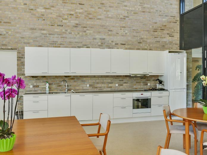 Større, lyst køkken op ad murstensvæg. Køkkenet har en ovn, en vaske og et køleskab samt en kaffemaskine. Foran køkkenet er der borde og stole.