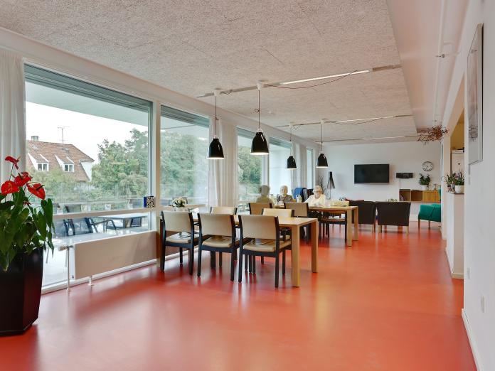 Fælles opholdsrum med bord, stole og grønne planter samt sofaområde med fjernsyn. Rummet lyst med store vinduespartier, hvide vægge og orange linoleumsgulve.