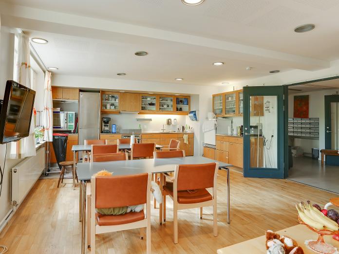 Fællesrum med stort køkken og to borde med stole. På væggen hænger et tv. 