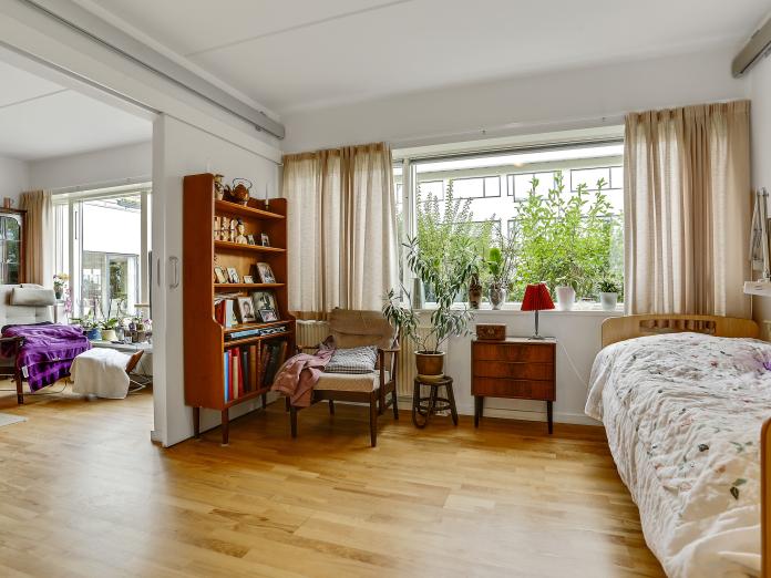 Soveværelse med seng, kommode, lænestol, reol og vindue med gardiner. Udenfor et par træer. Kig ind til stuen. Der er skydedør mellem de to rum.