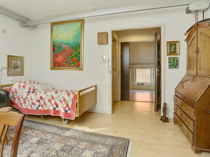 Lyst værelse med loftslift, plejeseng og personlige møbler som chatol, lænestol og gulvtæppe. 