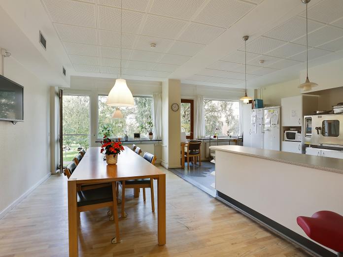 Fælles spiseområde med langbord og stole samt en serveringsdisk og et åbent køkken. 