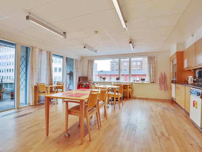 Lyst fællesrum med større køkken, borde og stole i lyst træ samt direkte udgang til fælles altan.