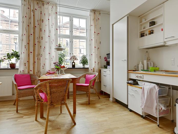 Lys stue med højt til loftet, store vinduer samt tekøkken med indbyggede skabe, vask og køleskab. Stuen er desuden indrettet med personlige møbler og potteplanter i vinduerne.