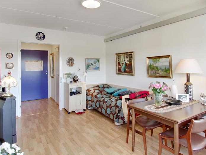 Lyst værelse indrettet med plejeseng og loftslift samt personlige møbler som spisebord, stole og fjernsyn.