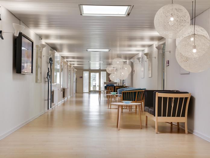 Lang, lys gang med hvide vægge og lofter og ovenlysvinduer samt sofaområder med fjernsyn og hyggelige lamper.