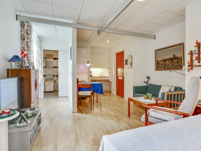 Stor, lyst værelse med loftslift og tekøkken. Værelset er indrettet med personlige møbler som seng, sofa, sofabord og lænestol samt lille spisebord med stole.