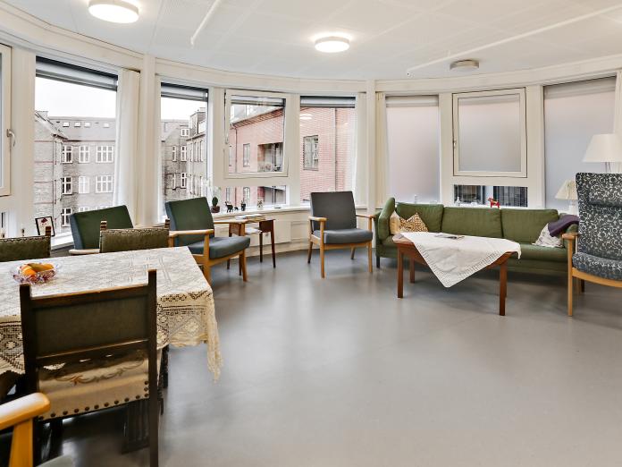 Halvrund stue med store vinduespartier. Stuen er indrettet med lænestole, sofaer og sofabord samt et bord med stole omkring. Bordene er dækket med duge, og møblerne er af ældre dato.