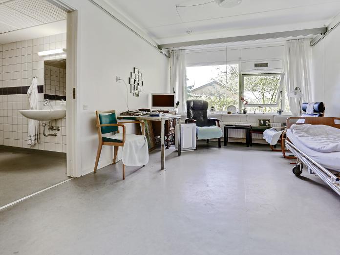 Lyst værelse med stort vinduesparti. Værelset er indrettet med plejeseng, loftslift, bord og stol samt lænestole og sofabord. Fra værelset er der adgang til et flisebadeværelse.