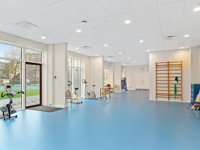 Stort motionslokale med hvide vægge, blåt linoleumsgulv og store vinduespartier ud mod grønt område. Rummet er indrettet med ribber, kondicykler og andre træningsredskaber. 