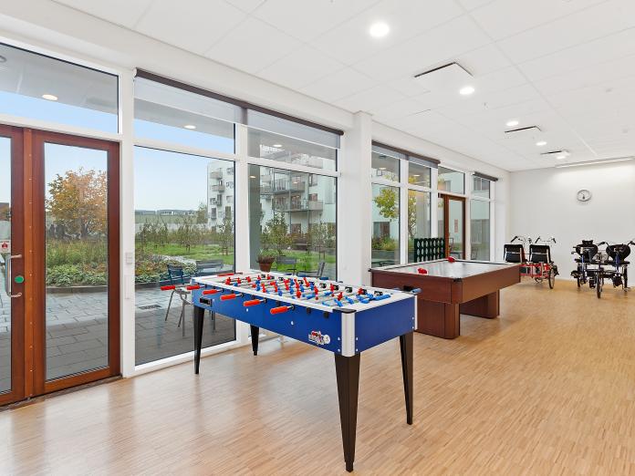 Fællesområde med store vinduespartier ud med gårdareal. Rummet indeholder et airhockeybord, et bordfodboldbord og to twinbikes. 