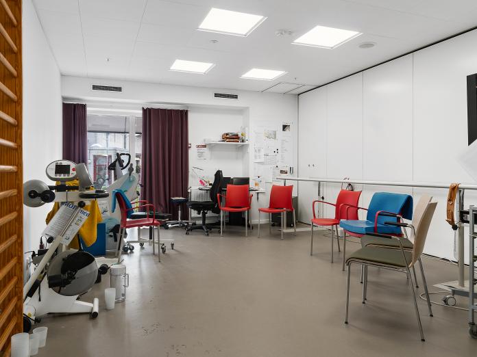 Lyst motionsrum med træningsmaskiner, ribbe, barre og stole stillet i en halvcirkel.