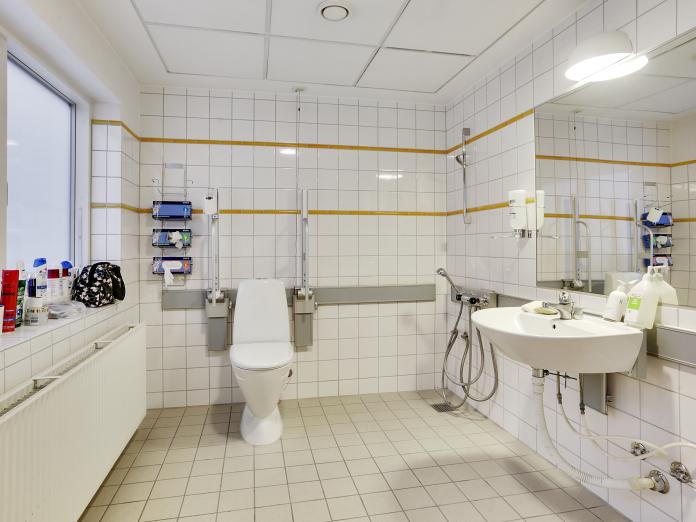 Stort, lyst badeværelse i hvide klinker med toilet, vask, stort spejl og bruser.  