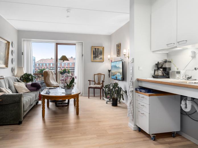 Lys stue med gulv til loft-vinduer og adgang til altan samt hvidt tekøkken. Stuen er møbleret med personlige møbler, fjernsyn og billeder på væggene.