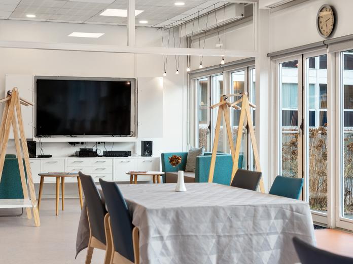 Stort, lyst fællesrum med terrassedøre ud til gårdhave. Rummet er indrettet med borde og stole, garderobestænger og et stort fjernsyn på endevæggen. 