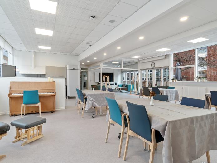 Stort fællesrum med store vinduer og terrassedøre. Rummet har også et køkken og er indrettet med borde, stole, klaver og en lænestol.