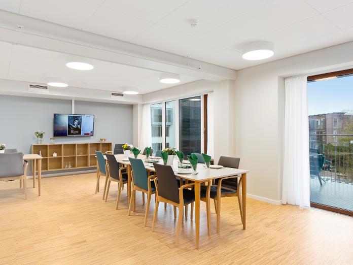 Fællesrum med langbord med stole omkring, fjernsyn og reoler på endevæggen samt store vinduespartier ud med altan. 