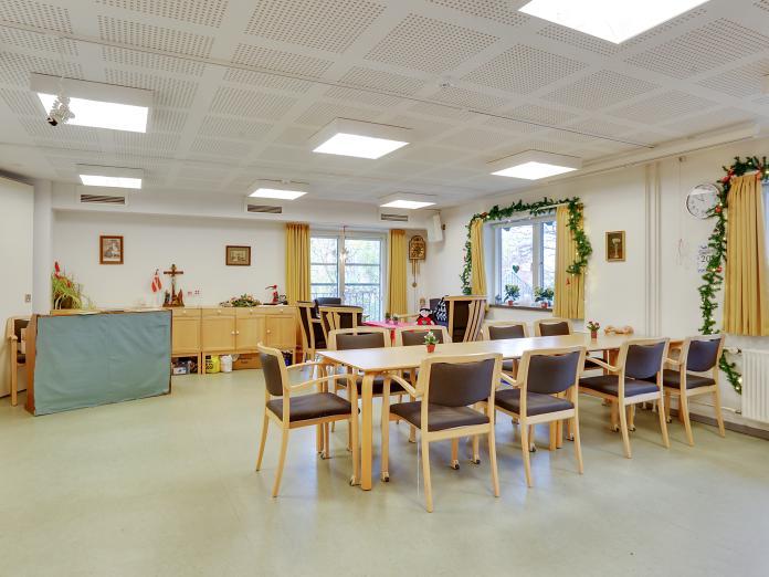 Fællesareal med klaver, langbord og stole, hyggekrog med lænestole og sofabord samt grønne planter og nipsgenstande. 