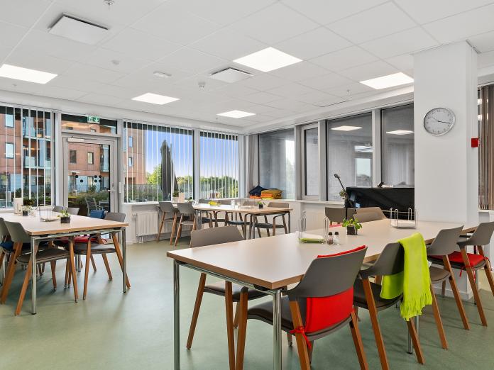 Lyst fællesrum med store vinduespartier. Rummet er blandt andet indrettet med langborde, stole og et klaver.