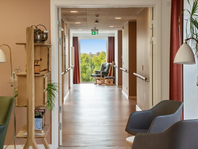 Kig ned ad lang gang med gelænder og gulv til loft-vindue i endevæggen, hvorfra man kan se grønne træer.