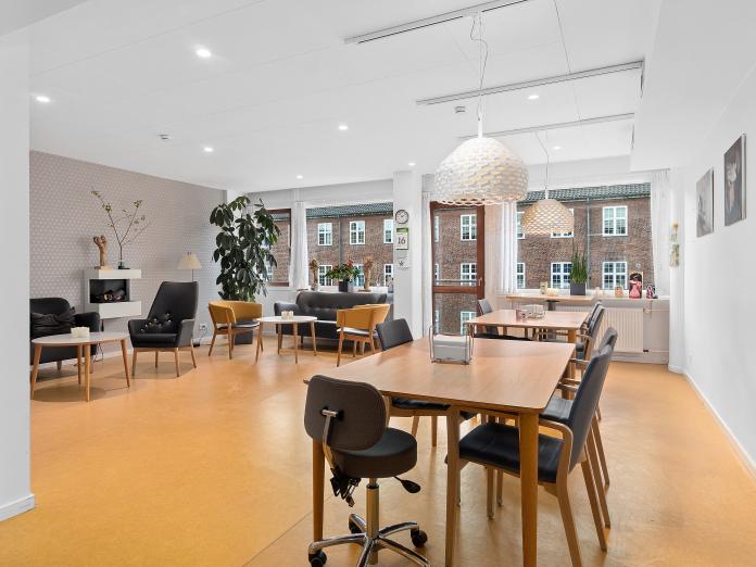 Fællesrum med store vinduespartier og altandør. Rummet er indrettet med to borde med stole omkring samt sofa, lænestole, sofaborde og en elektrisk pejs.