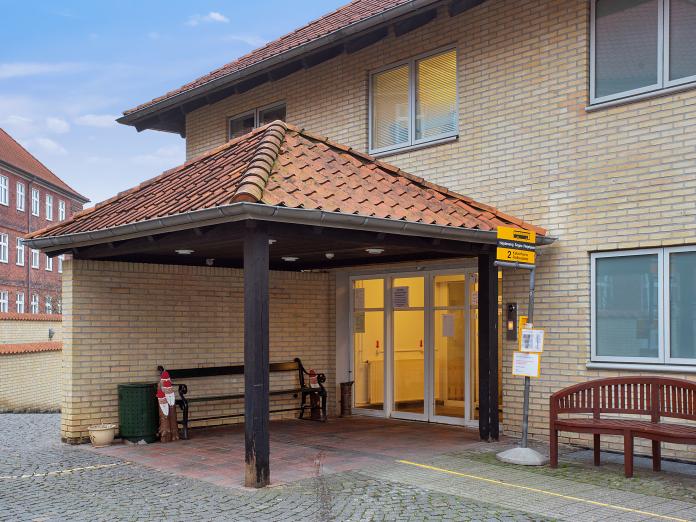 Indgangsparti til gul murstensbygning med røde tegl. Ved indgangen er placeret to bænke og et busstoppeskilt.
