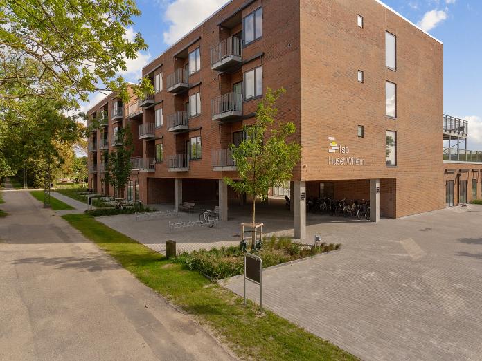 Moderne etagebygning i røde sten med stålaltaner og søljer i beton omkranset af fliser, grønne bede og grønne træer.