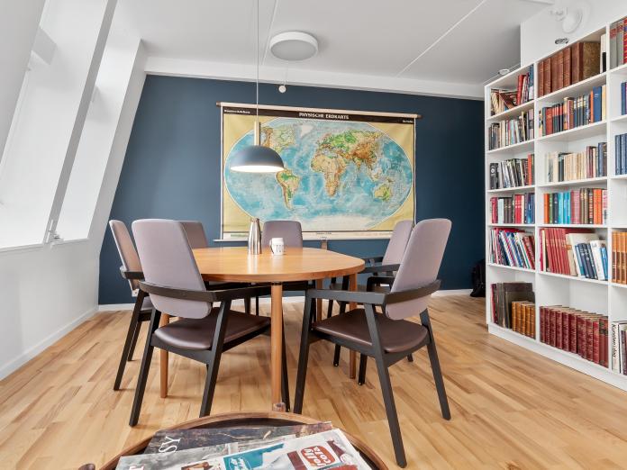 Fælles rum indrettet som bibliotek med reoler fylde med bøger, et ovalt bord med stole omkring samt et bakkebord med aviser og blade. På væggen hænger et skolekort over verden.