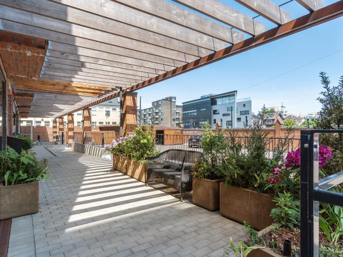 Fælles fliseterrasse med pergola. Terrassen er indrettet med kurvesofa og store plantekasser med grønne planter og blomster.