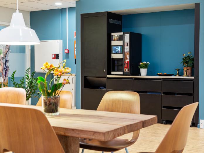 Fællesområde med tekøkken med kaffemaskine og køleskab. Rummet er indrettet med langbord og stole, blomster og grønne planter amt en magasinholder.